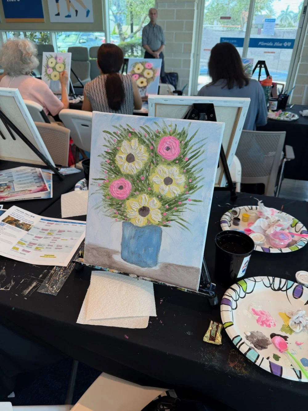 A painting of a flower bouquet with yellow daisies and pink roses in a blue vase, placed on an easel at a painting class.
