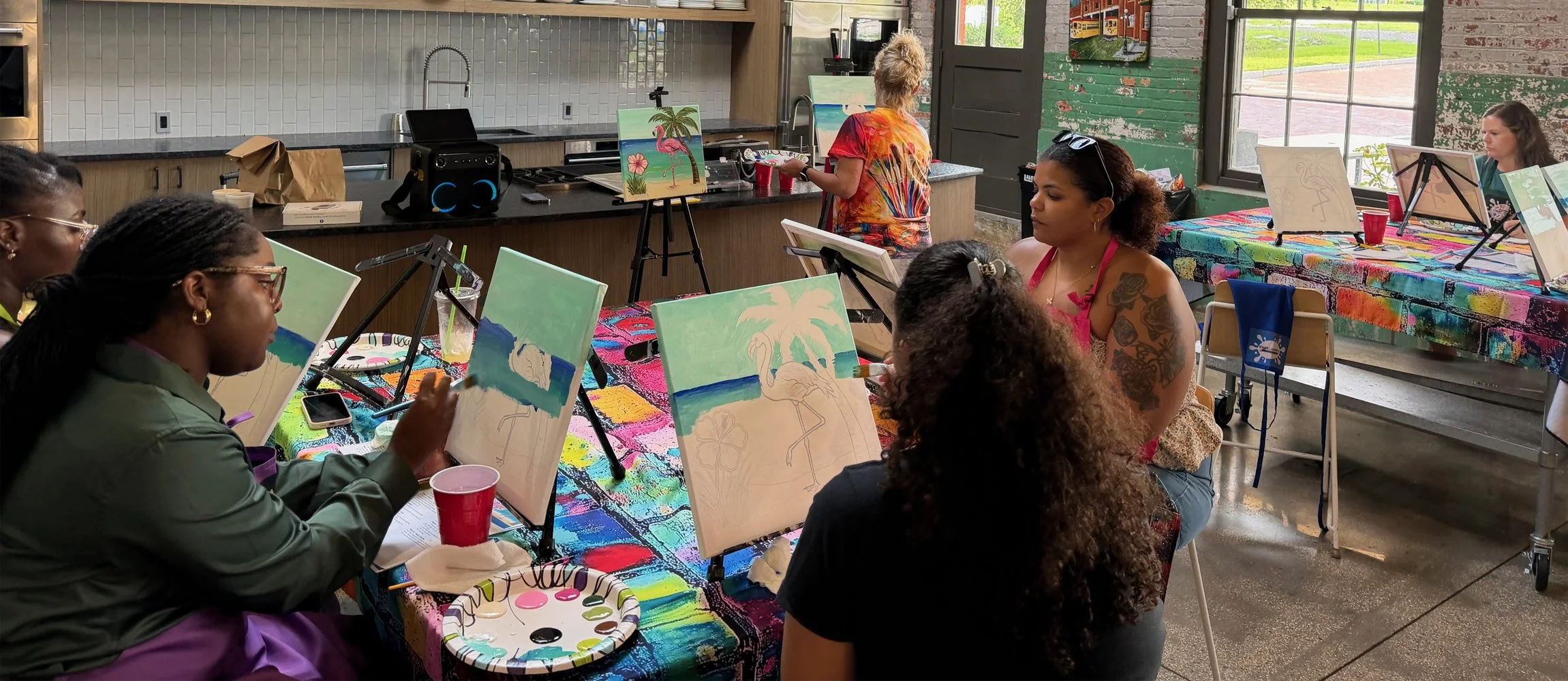 A group of women participating in a painting class in a rustic, colorful studio with windows and a kitchen area in the background. They are working on tropical-themed paintings of flamingos and palm trees.