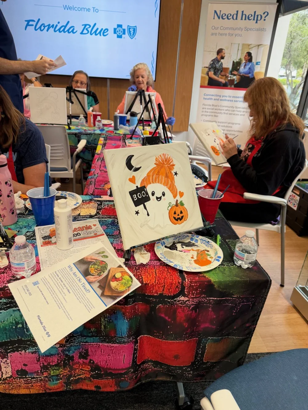 A group of people participating in a Halloween-themed painting activity, with a colorful tablecloth, water bottles, brushes, and Halloween artwork, including a ghost with an orange hat and pumpkin, on display.