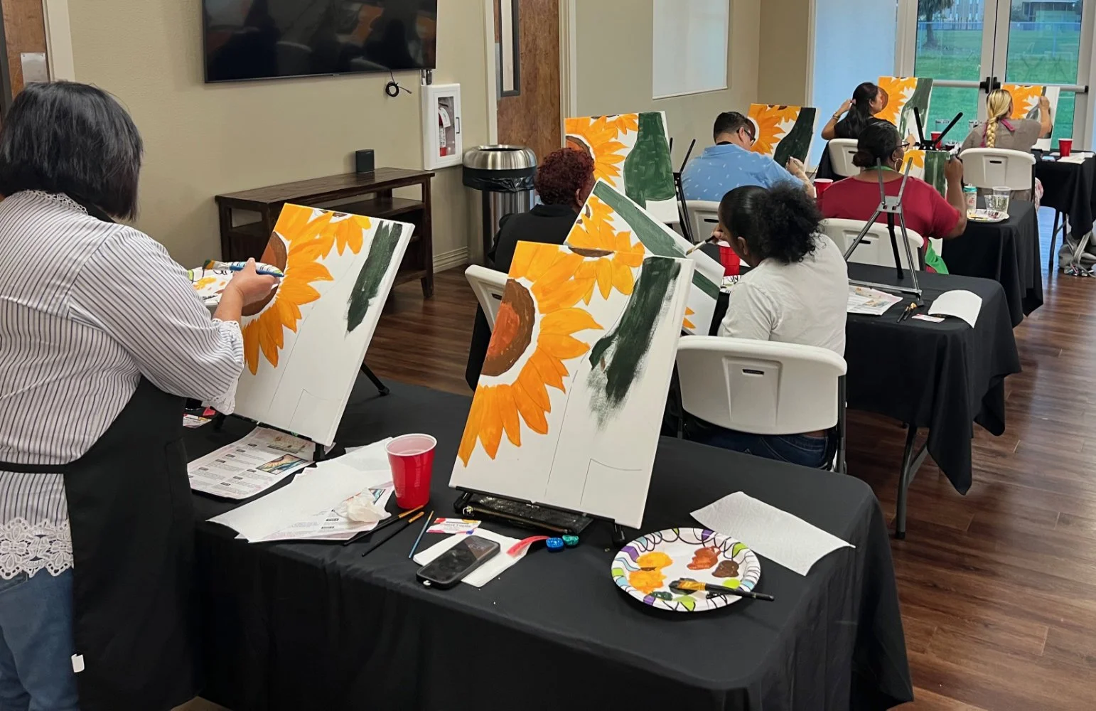 People painting sunflower artwork during an art class in a bright, well-lit room with large windows.