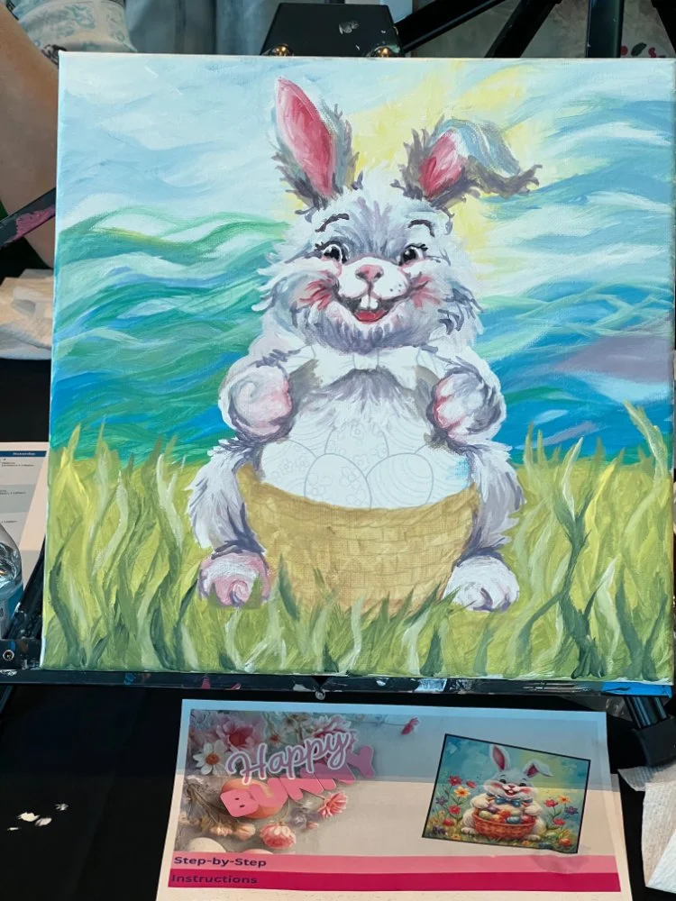 A colorful painting of a happy, animated bunny with pink inside its ears, sitting on green grass holding a basket of decorated eggs, with a bright sky and clouds in the background.
