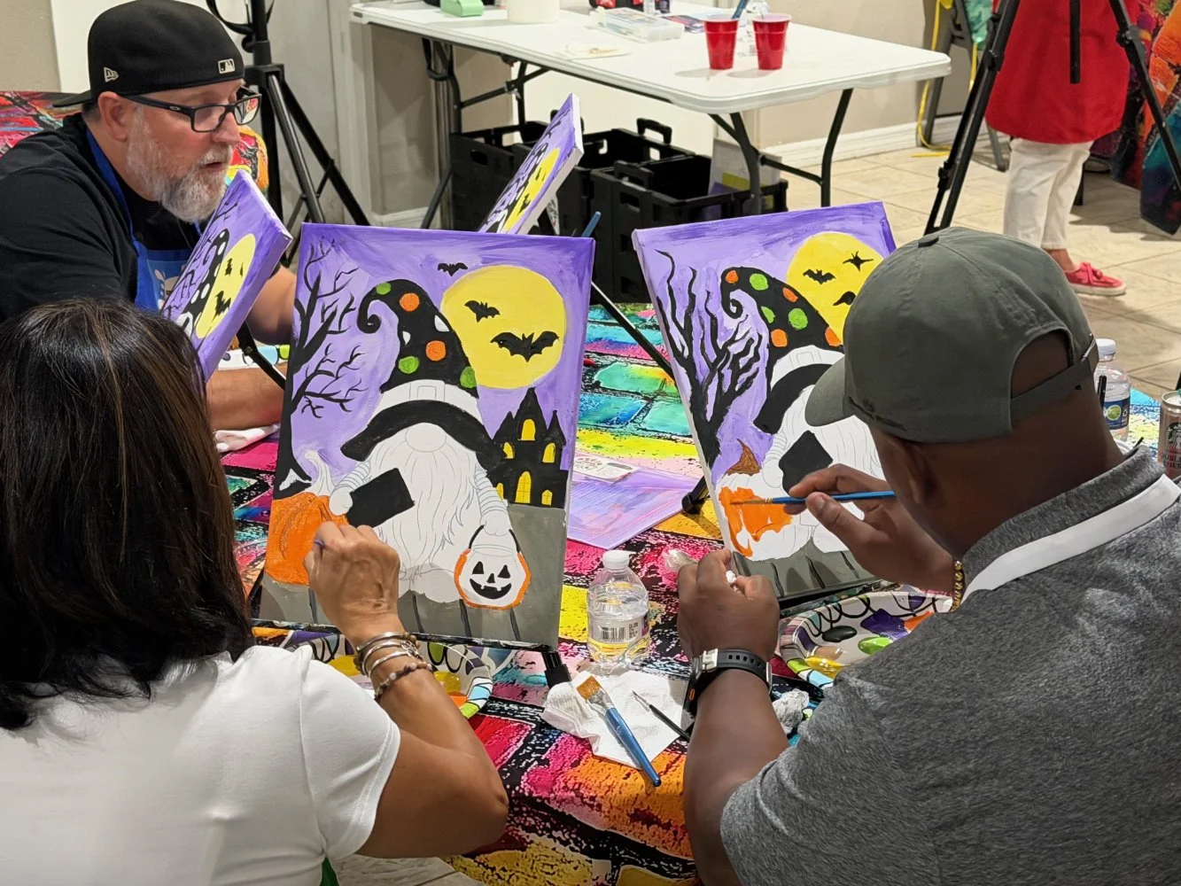 People painting Halloween-themed pictures of a witch with a purple background, full moon, bats, and trees at a table.