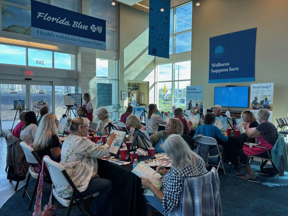 A group of people participating in an indoor art class with paintings and easels, in a brightly lit room with large windows and posters about wellness and health.