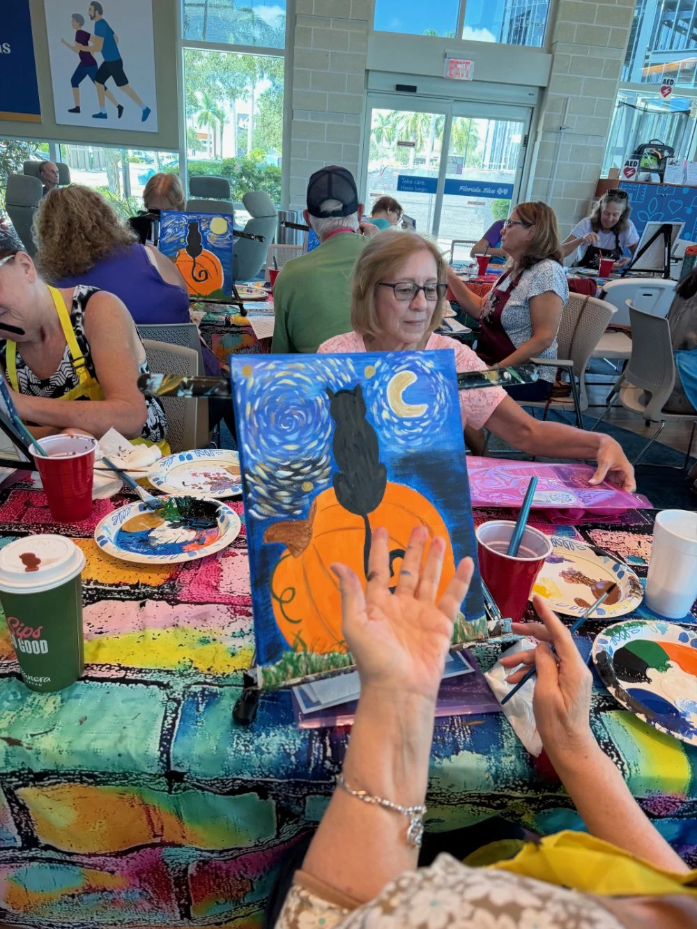 A group of people participating in a painting class, creating artworks inspired by Vincent van Gogh's style. The main focus is on a woman painting a Halloween-themed scene with a black cat, pumpkin, and night sky. The table is covered with colorful p