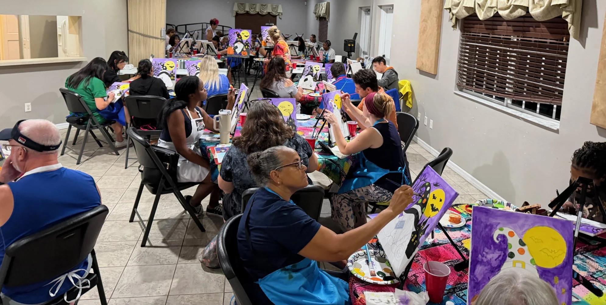 Group of people participating in a Halloween-themed painting event, creating spooky illustrations of bats, ghosts, and a haunted house, with a decorated table and paintings featuring a full moon, trees, and Halloween motifs.