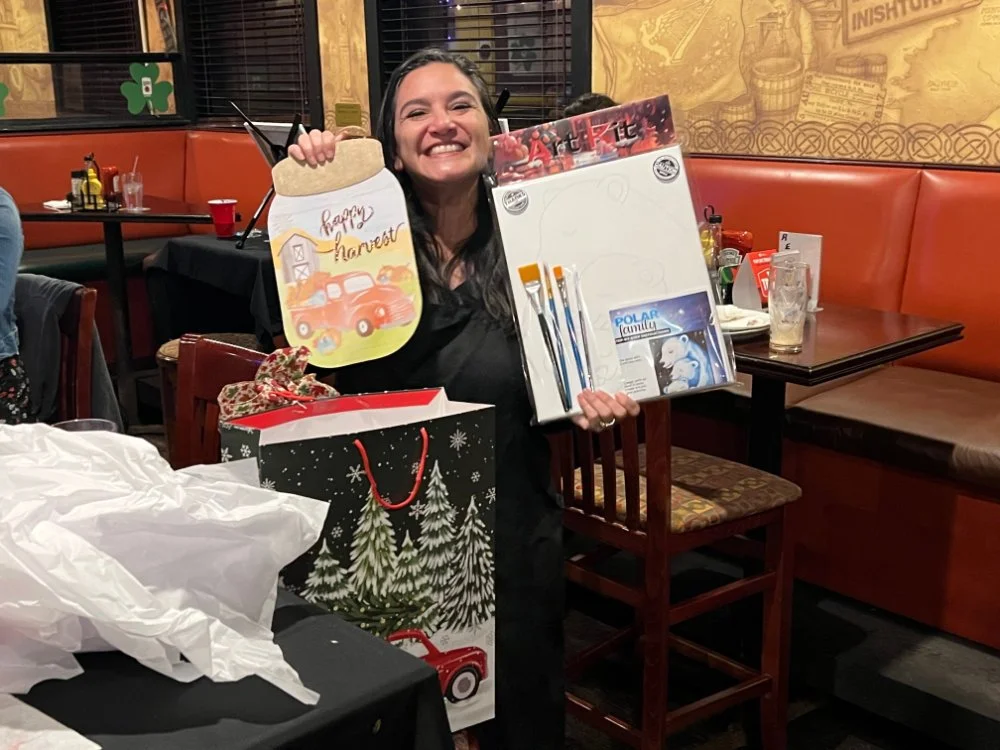 A woman smiling and holding Christmas gift bags and a snow-themed drawing pad in a restaurant.
