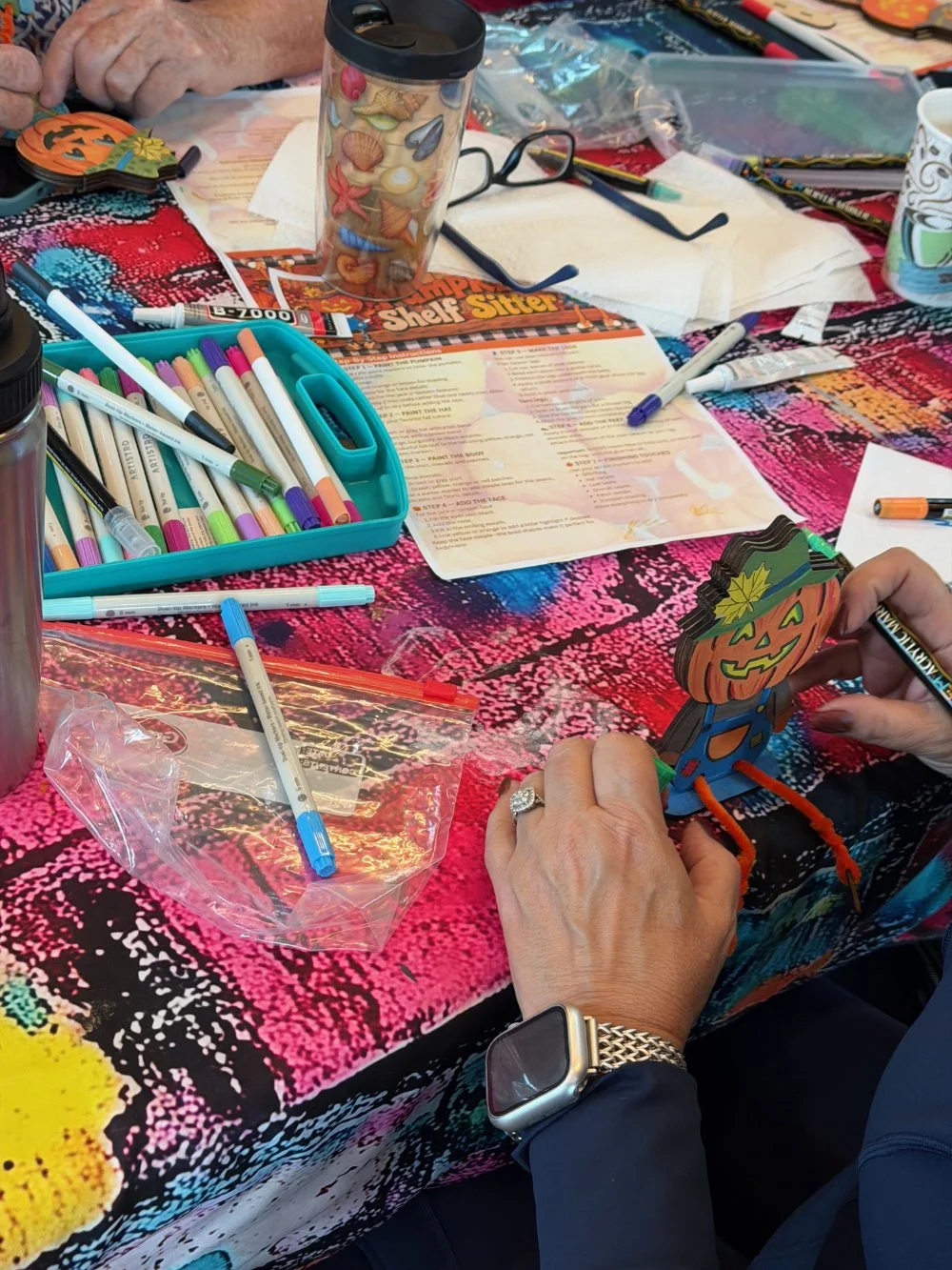 People crafting Halloween paper decorations at a table with colorful markers, scissors, and printouts.
