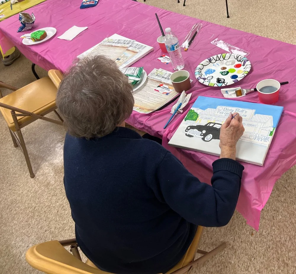 An elderly woman sitting at a table with a pink tablecloth, painting a picture of a car in front of a painting of a car that says 'Dirty Bill's Ice Cream' at a craft or painting event.