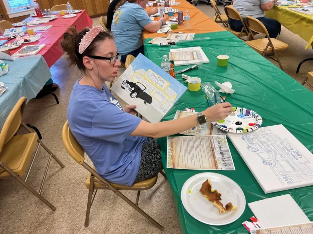 A woman with glasses and a headband sitting at a decorated table, painting a picture of a school bus with a black car on a paper plate, with various art supplies and a slice of pizza on her table in a social gathering setting.
