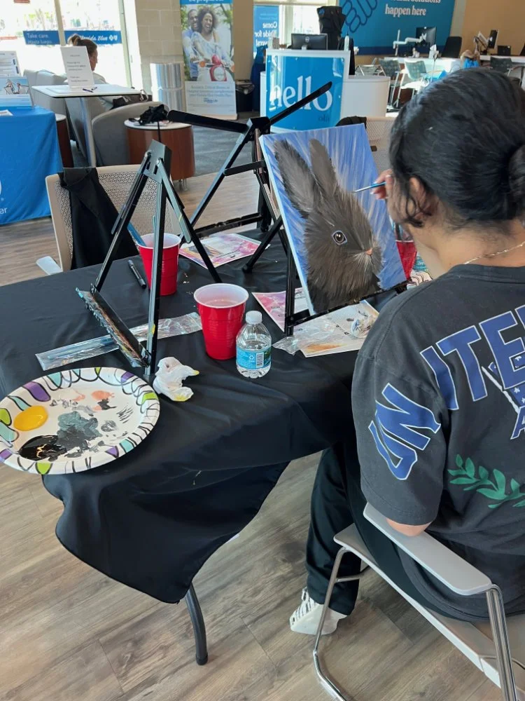 A person painting a realistic depiction of a brown rabbit on a canvas at an indoor art event. The artist is seated at a table with painting supplies, a red cup, a water bottle, and a paper plate with paint. In the background, there are informational 