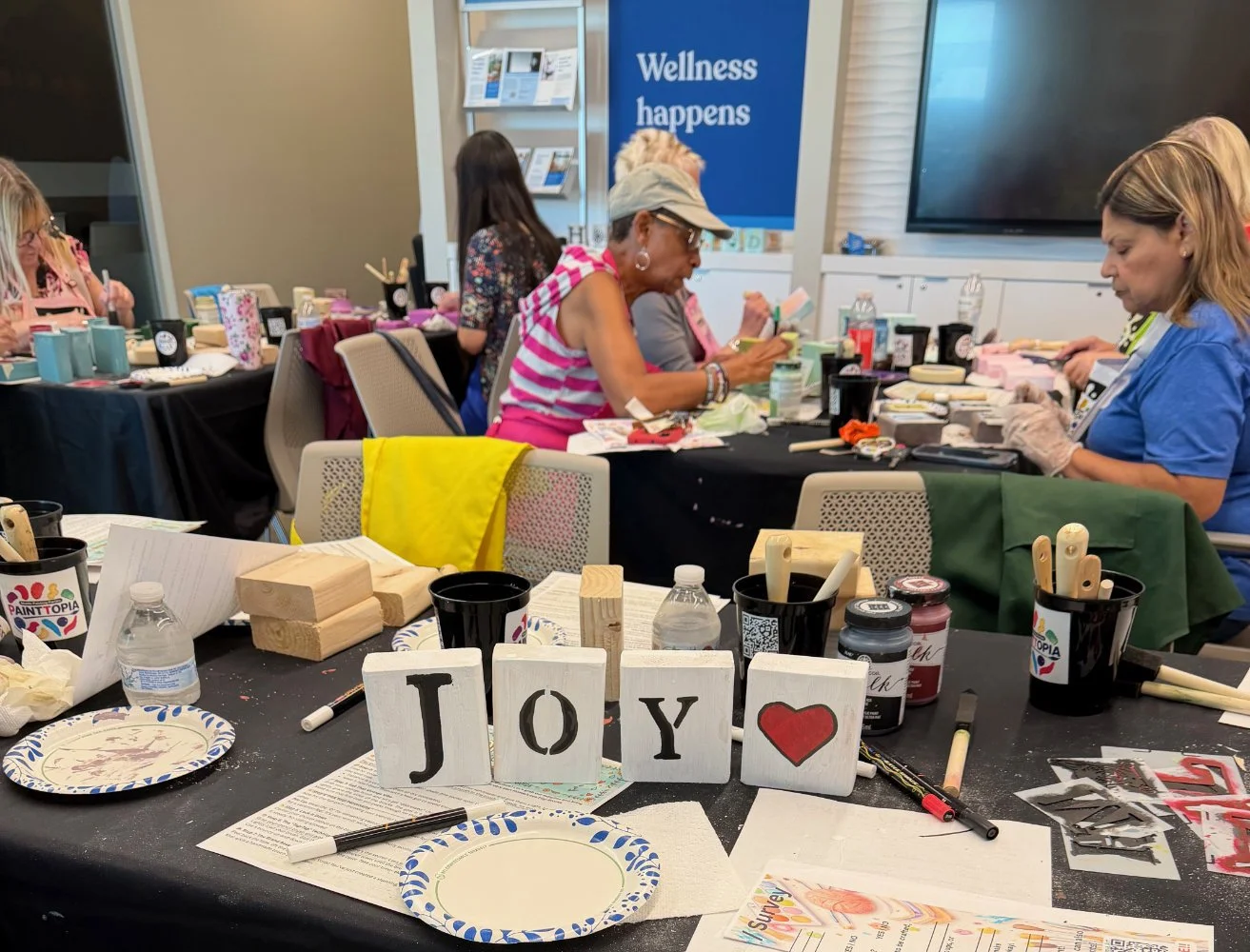 People participating in a crafting workshop at a table with paint, brushes, and wood blocks, with a sign that says 'Joy' on the table.