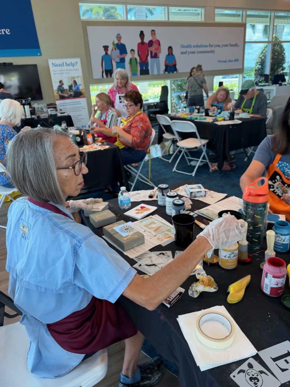 A group of people, mostly women, participating in a craft activity at a community event inside a room with large windows. One woman in the foreground is working with art supplies, including paints, brushes, and paper, and appears focused on her proje