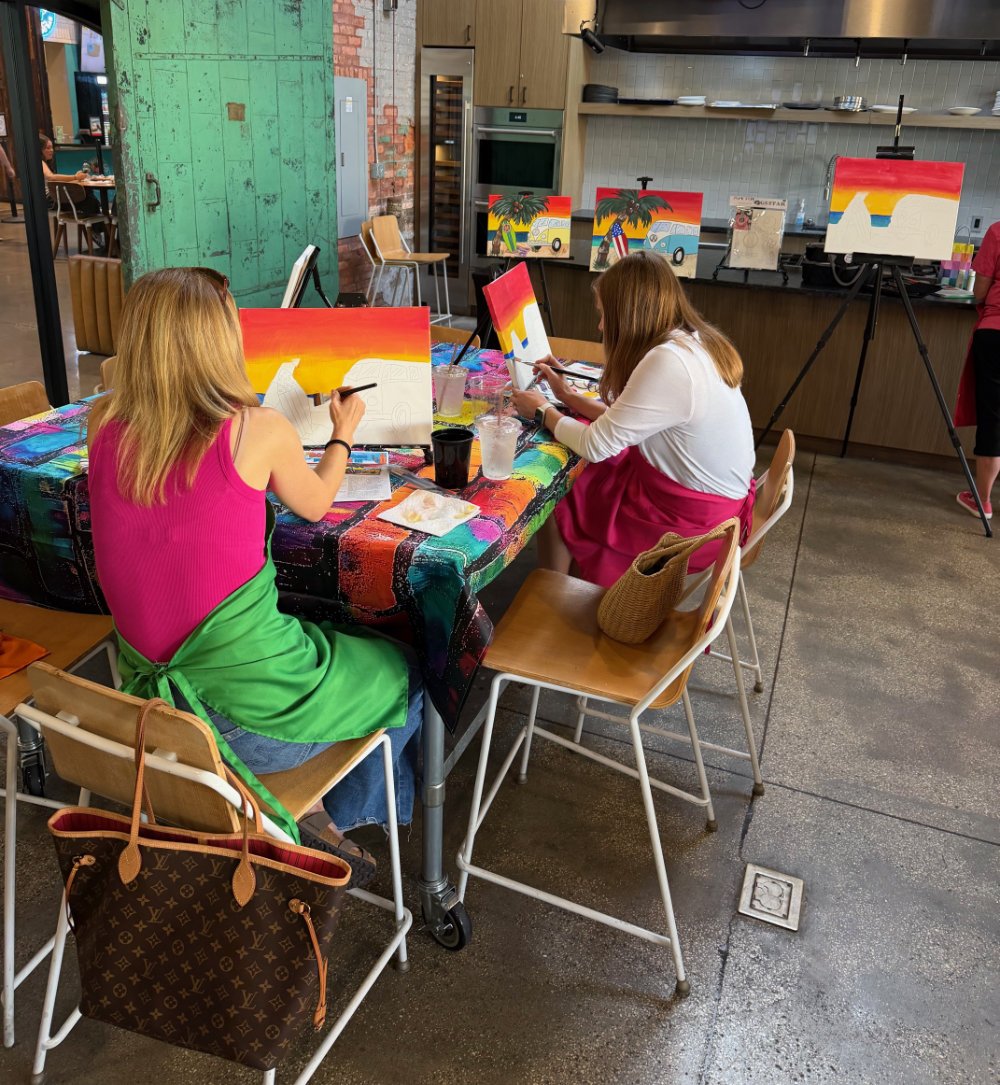 Two women painting with tropical and sunset themes on canvases at a table in an art studio or cafe, with other paintings displayed in the background.
