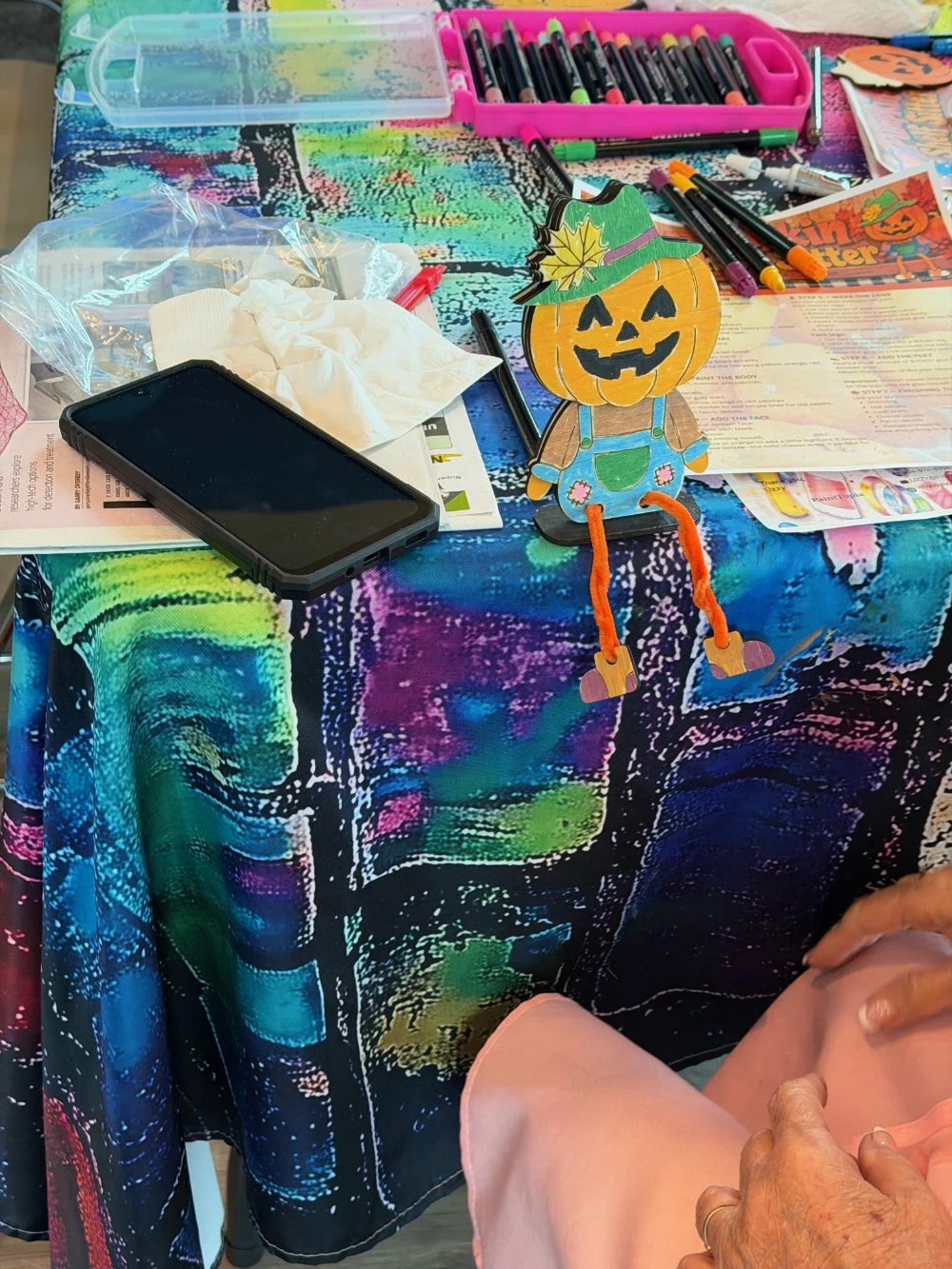 A table decorated with a colorful, abstract tablecloth holds various arts and crafts supplies, including markers, a smartphone, tissues, and a paper pumpkin with a haunted hat and pumpkin face.