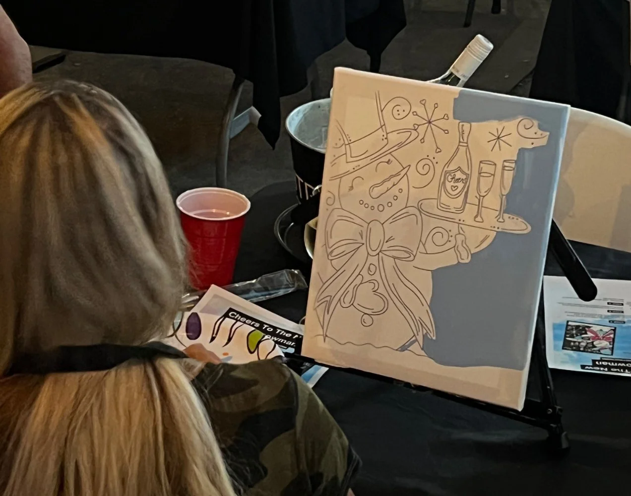 A woman with long blonde hair observing a canvas with a line drawing of a festive party scene, including a champagne bottle, glasses, and ribbons, beside a red plastic cup on a table at an indoor event.