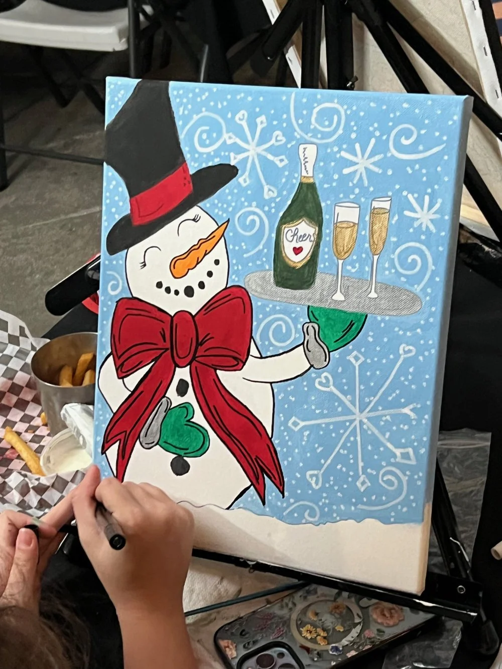 A person painting a festive holiday scene on a canvas featuring a snowman wearing a black top hat with a red band and a red bow tie, holding a tray with a bottle labeled 'Cheers' and three glasses of champagne, with snowflakes and swirls in the backg