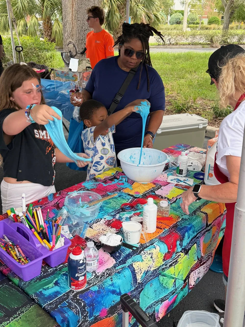 Children and adults making slime at a colorful table outdoors.