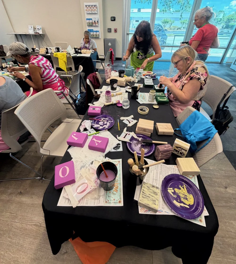A group of women engaged in a craft workshop around a table filled with wooden blocks, paint, brushes, and decorative items, in a well-lit indoor space with large windows and a view outside.