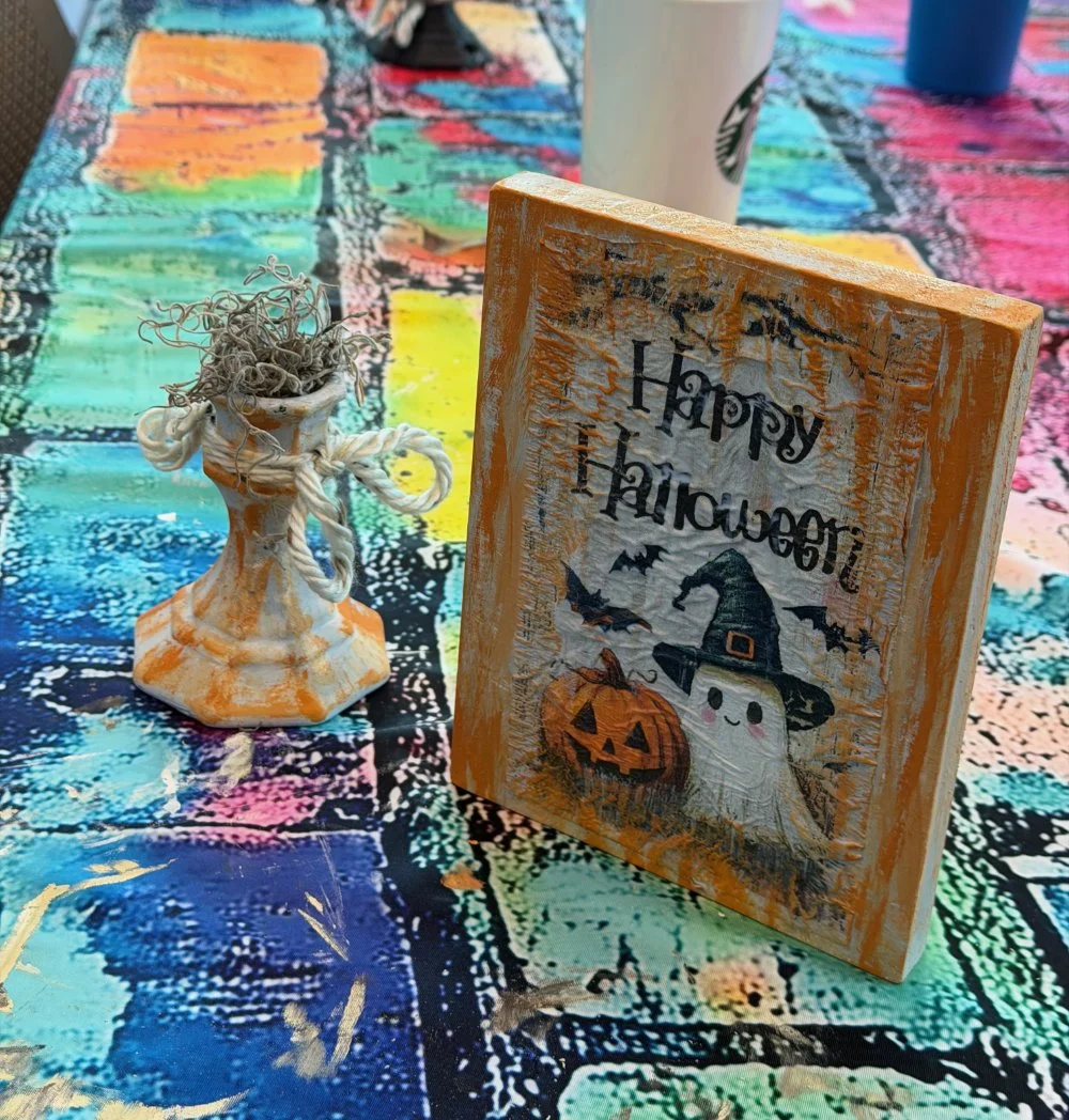 A Halloween-themed table decoration setup featuring a painted wooden sign with a smiling ghost wearing a witch's hat and a carved pumpkin, next to a small ceramic vase with curly twigs. The background consists of a colorful, abstract tablecloth with 