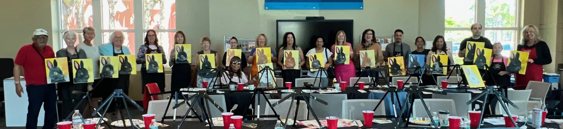 Group of people standing in a room holding up paintings of rabbits. There are tables with red cups and art supplies, cameras on tripods, and large windows in the background.