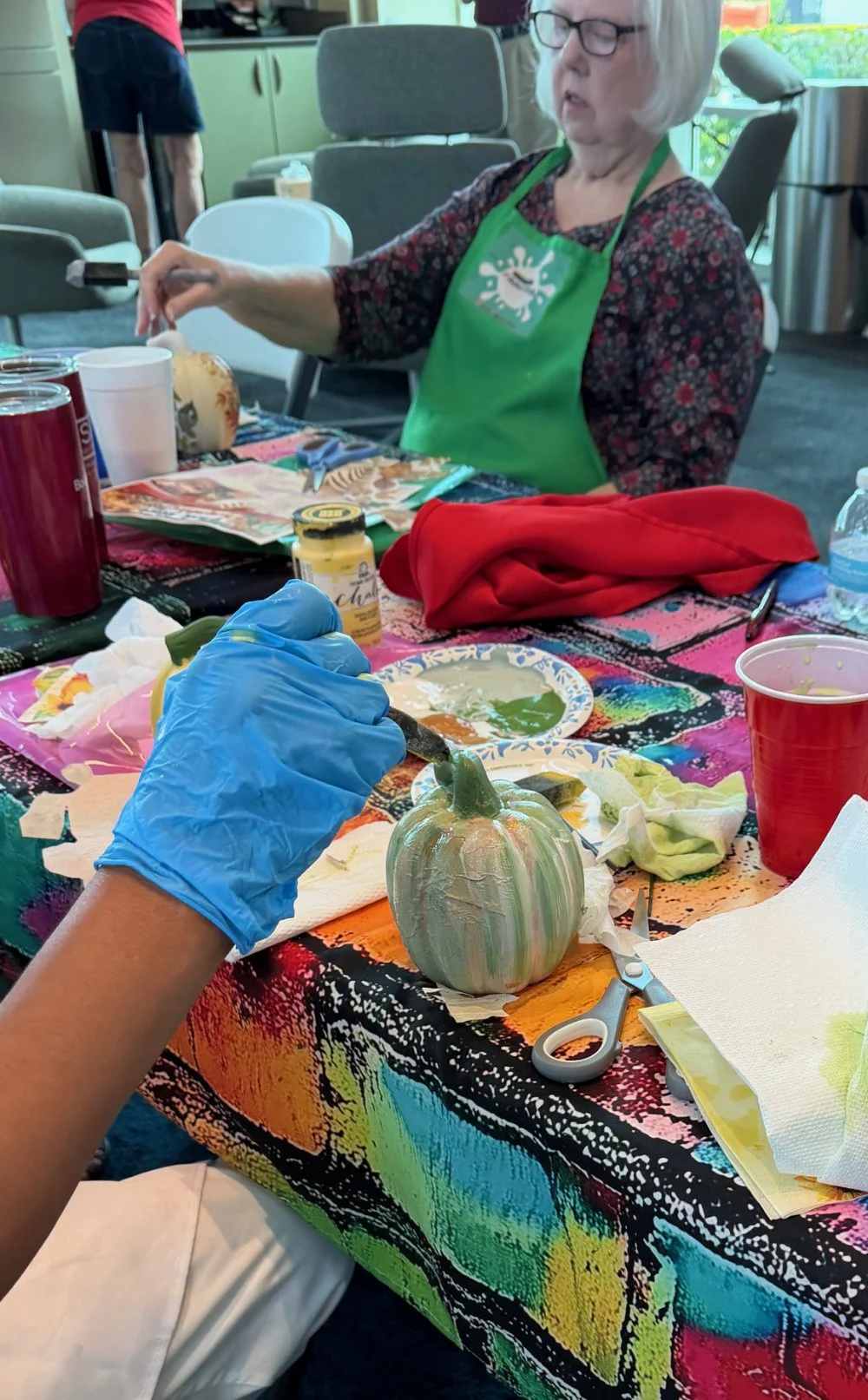 A woman with gray hair and glasses is sitting at a table decorating a pumpkin with paint. The table is covered with a colorful cloth, and another person wearing a blue glove is also painting the pumpkin. Various art supplies, cups, and paper towels a