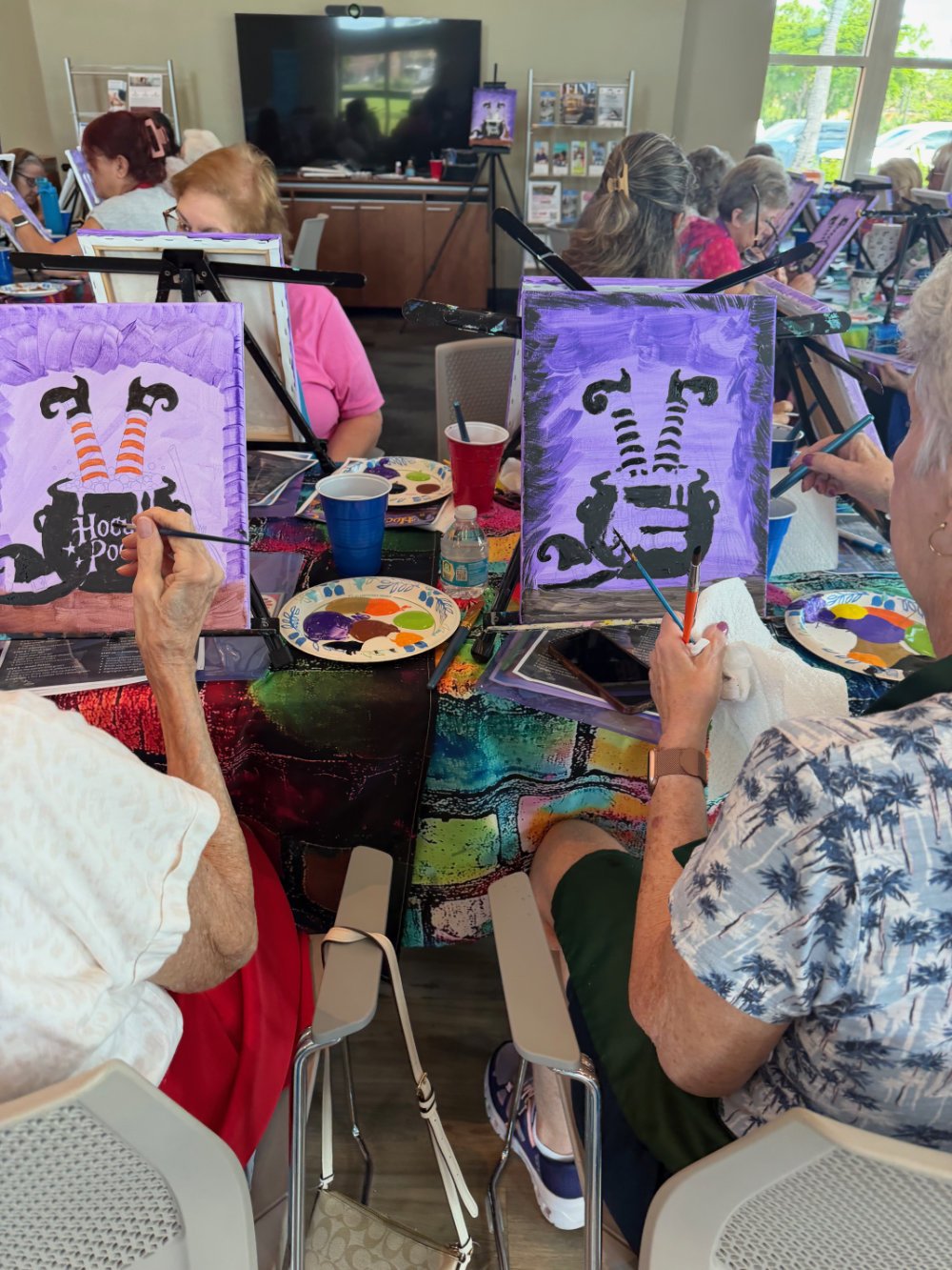 Older adults participating in a group painting class, creating artwork of a black cat upside down with orange striped shoes and a purple background.