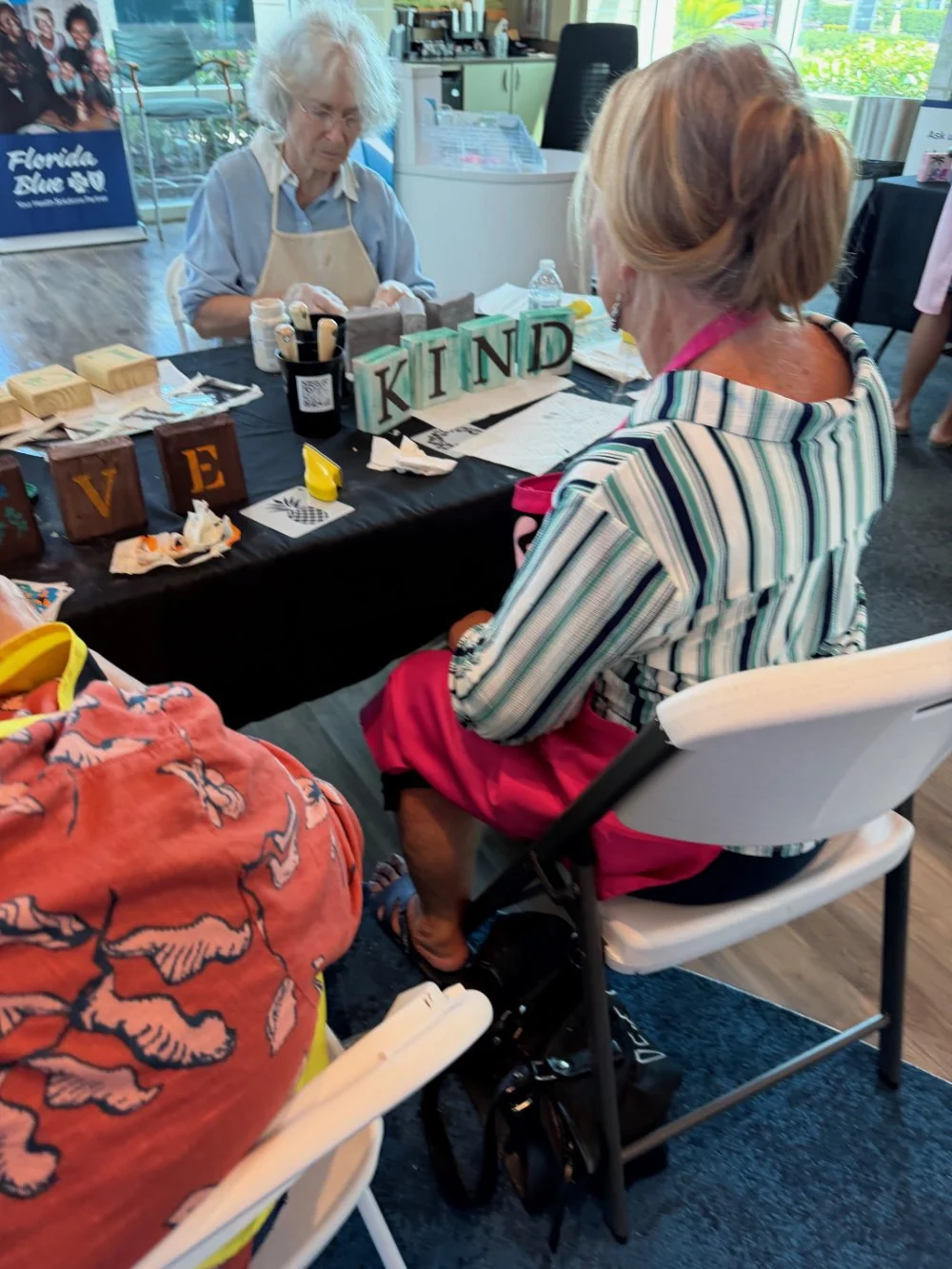 A woman sitting at a table at a craft or art event, talking to an elderly woman with white hair behind the table, which displays blocks spelling "LOVE" and "KIND" and various craft supplies. The scene is indoors with large windows in the background.