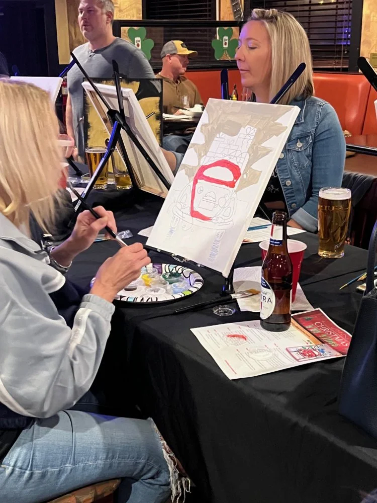 A woman is painting on a canvas at a table in a restaurant or bar. Other people are seated and standing around, with drinks on the table, and Christmas-themed decorations are visible in the background.