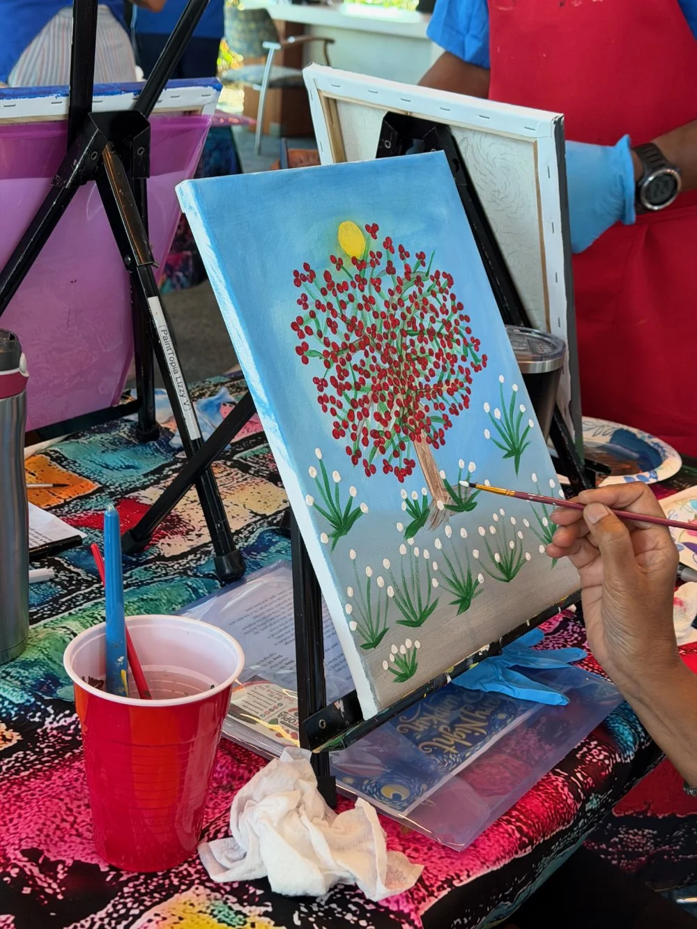 Person painting a colorful tree with red leaves and green grass on a small canvas at an outdoor art event, with various painting supplies and other canvases around.