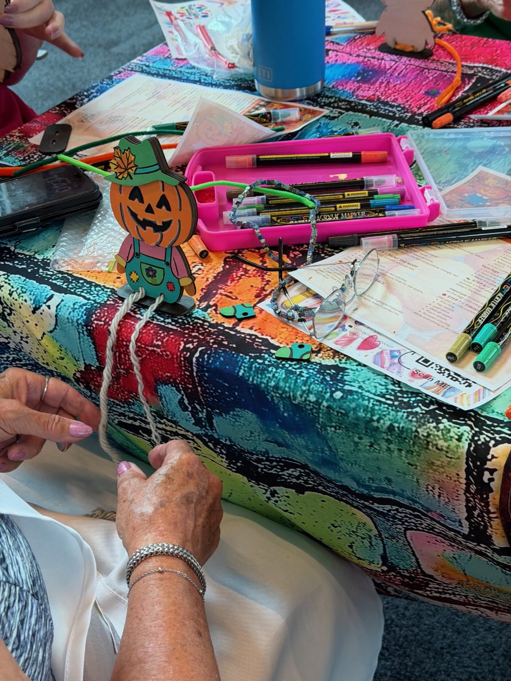 A colorful table with various art supplies including markers, pens, and paper. There is a decorative pumpkin figure with a hat and yarn legs. An elderly woman is working on a craft project, holding a white string.