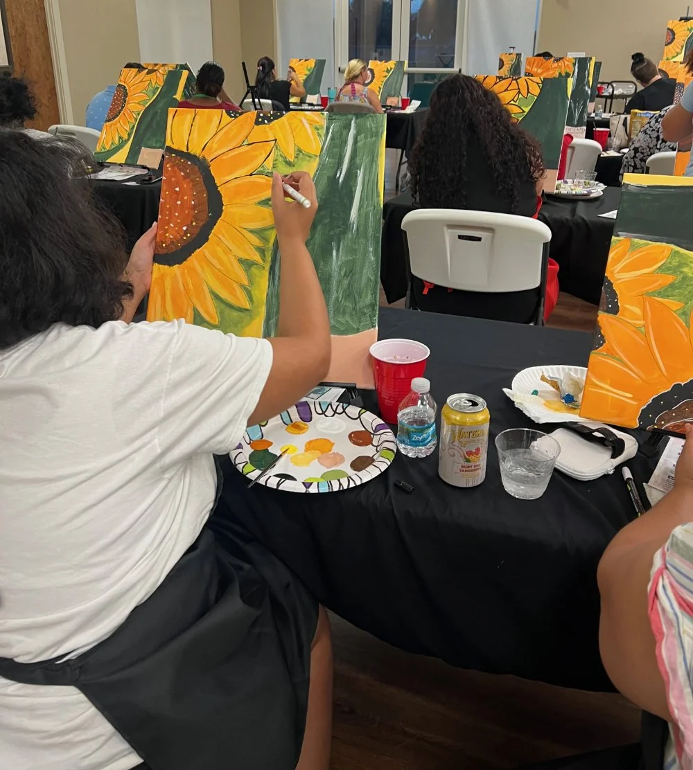 A group of people at an art class painting sunflower canvases. One person in the foreground is painting a sunflower with yellow petals and a brown center. The tables have painting supplies, drinks, and snacks.