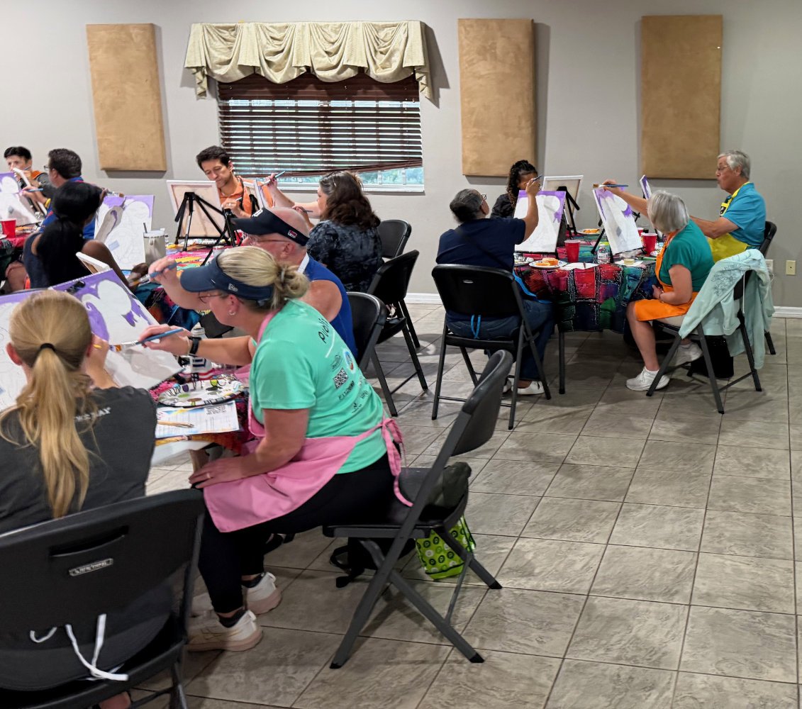 Group of people painting at tables in a room with beige acoustic panels and a window with a beige valance.