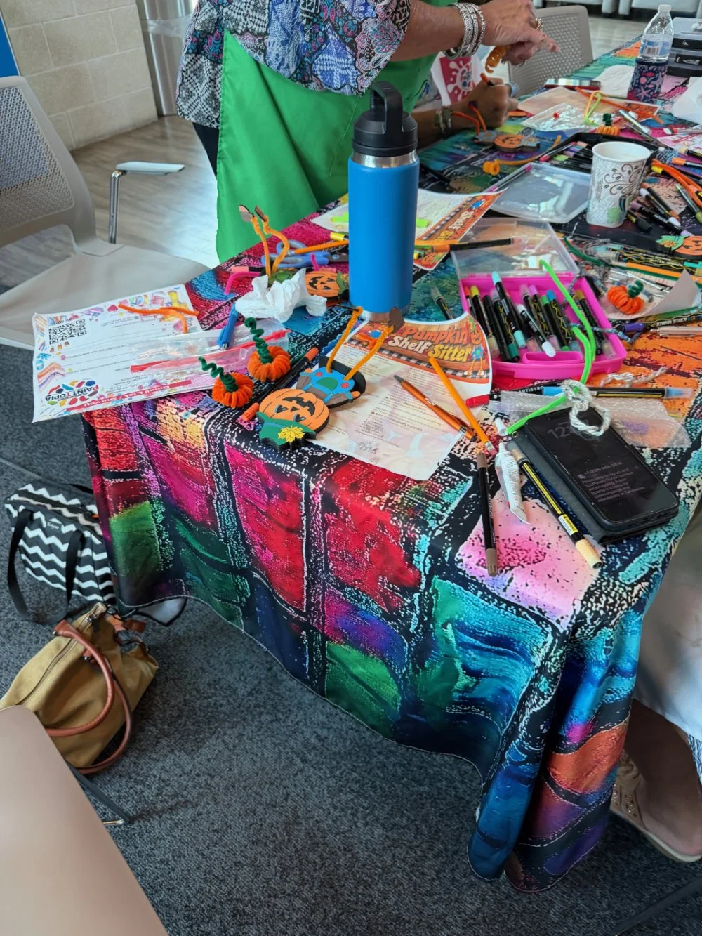 A colorful table covered with craft supplies, including markers, pipe cleaners, Halloween-themed decorations, a water bottle, and various papers, with people working on craft projects.