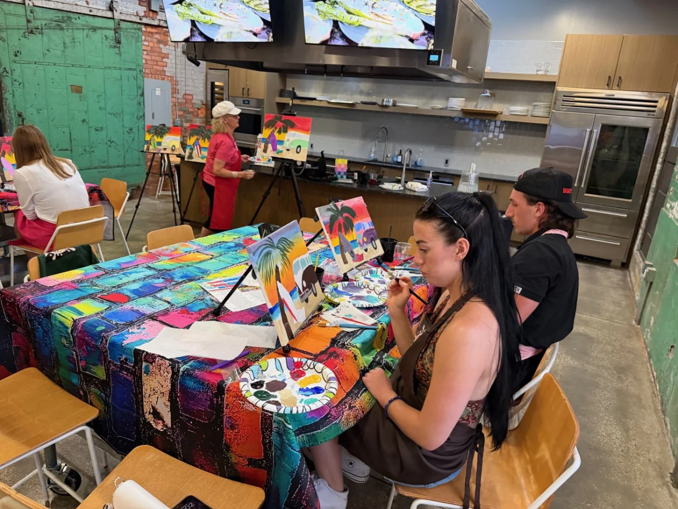 People painting tropical scenery on canvases at an indoor painting class with colorful artwork and supplies on a table.