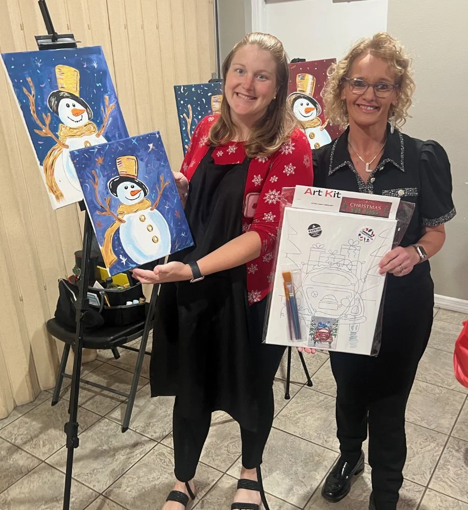 Two women smiling, holding a completed Christmas art project and a set of drawing supplies. Behind them, several finished paintings of snowmen are displayed on easels.