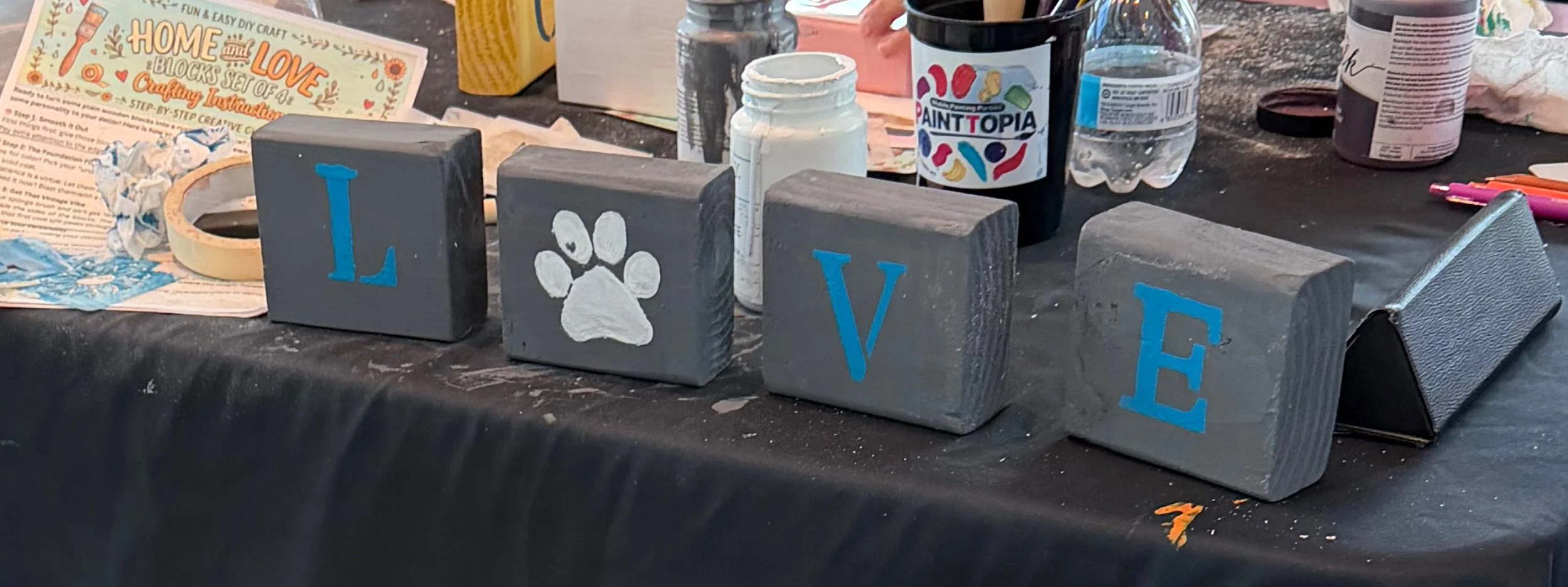 Decorative blocks spell out the word 'LOVE', with the letter 'O' replaced by a paw print. The blocks are painted gray with blue letters and are displayed on a black table surrounded by craft supplies.