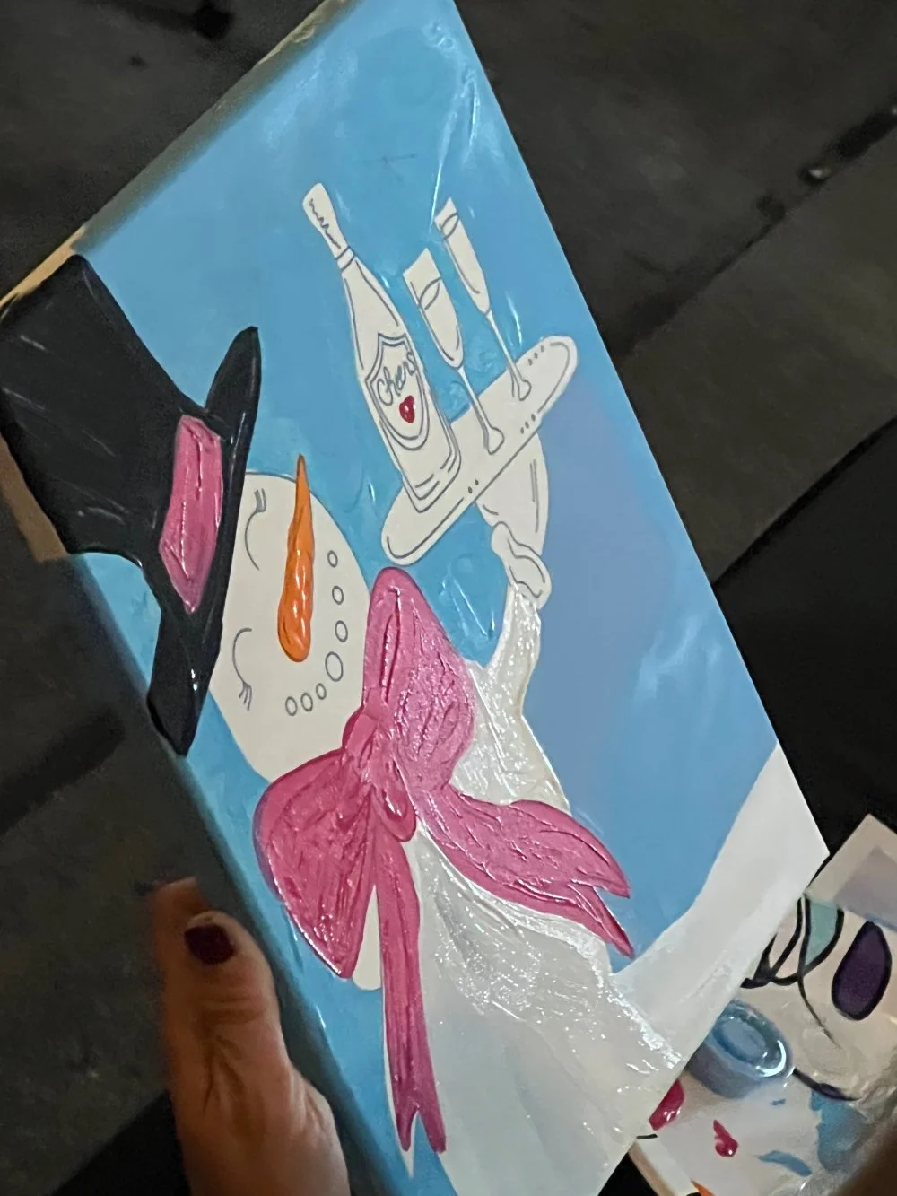 Colorful painting of a snowman with an orange carrot nose, pink bow, and black hat, with a tray of drinks and a bottle labeled 'Cheers' in the background.