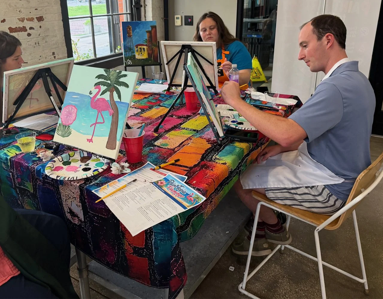 People painting tropical-themed pictures of flamingos, palm trees, and a beach scene at a group painting class.