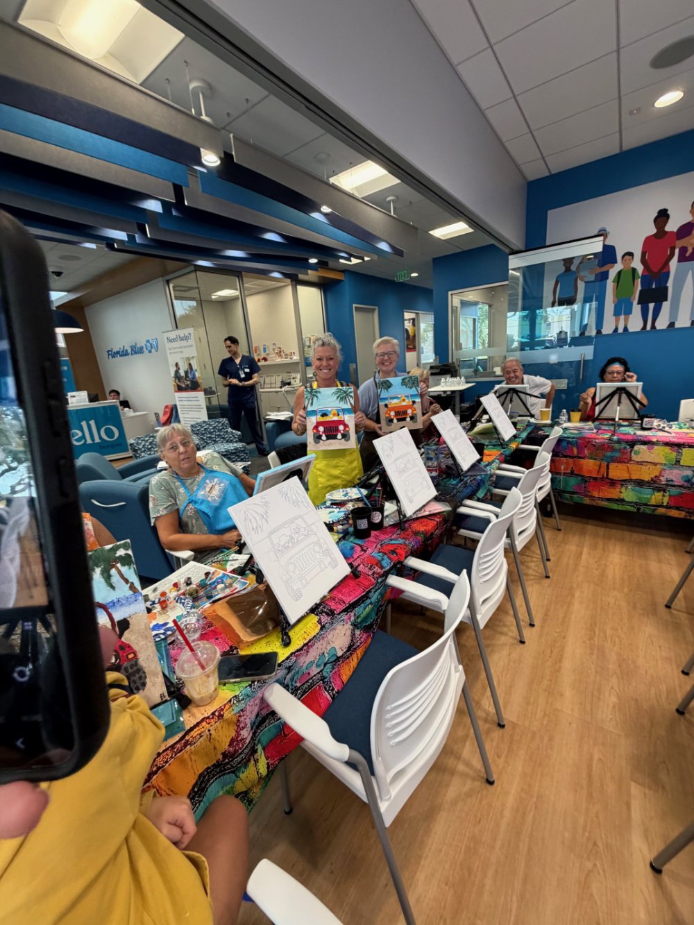 Group of people at an indoor painting class, smiling and holding canvases with colorful car paintings, seated at a long table with art supplies, in a cheerful, decorated room.