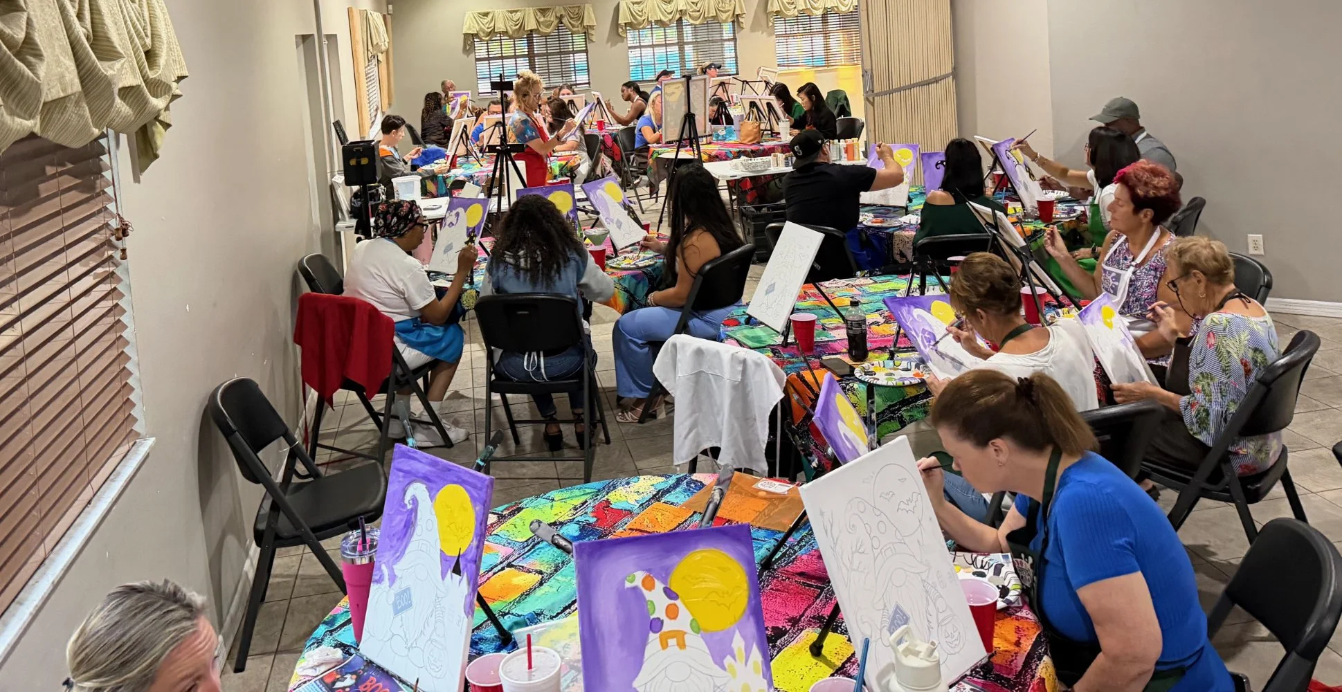 Group of people participating in a painting class, each working on their own canvas with colorful paintings, tables covered with vibrant tablecloths, and artwork depicting a moon.