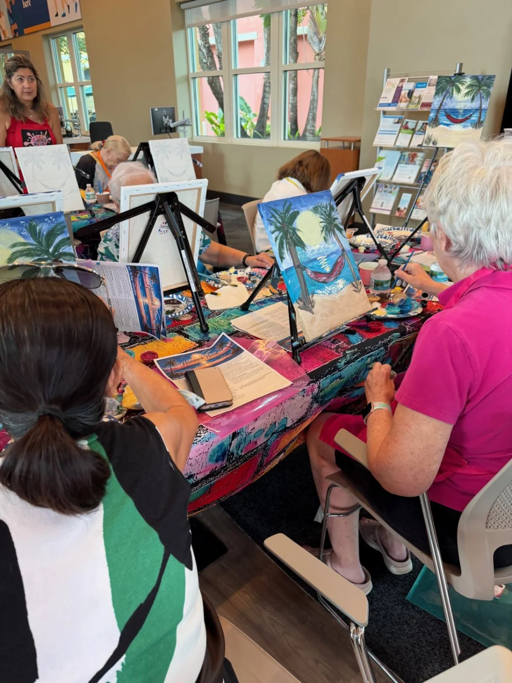 People participating in a group painting activity, working on tropical beach scenes with palm trees and a hammock, at a workshop in a brightly lit room with large windows and colorful tablecloths.