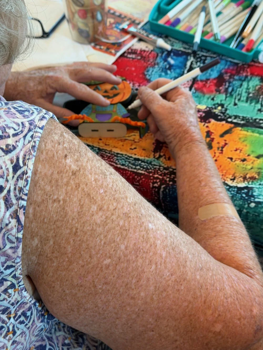 An elderly woman sitting at a colorful table, drawing on a small pumpkin-shaped object with a white marker. Her other hand is resting on the table, and there are various art supplies, such as markers, in a container nearby. She has a bandage on her a