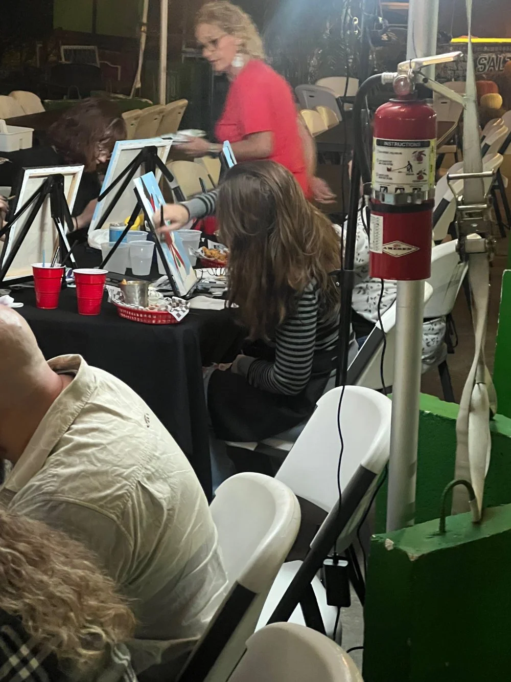 A group of people sitting at tables with disposable red cups, surrounded by painting supplies and easels, at an outdoor or semi-outdoor event. A fire extinguisher and some equipment are visible nearby.