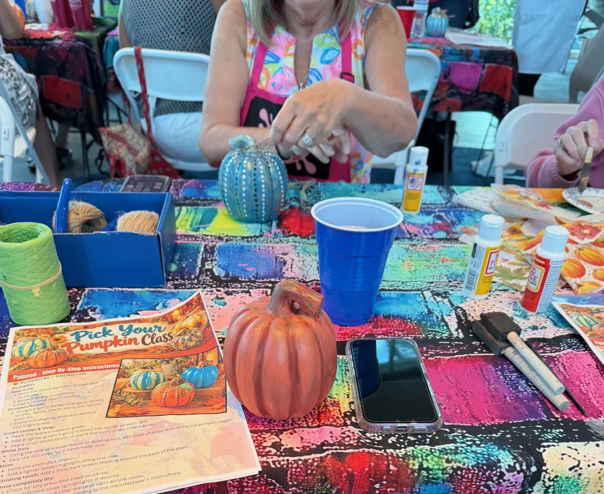 A woman at a table decorated with colorful tablecloths, working on painting a small pumpkin with blue and white accents. The table has various art supplies, a blue cup, a smartphone, and a printed pumpkin painting instruction sheet.