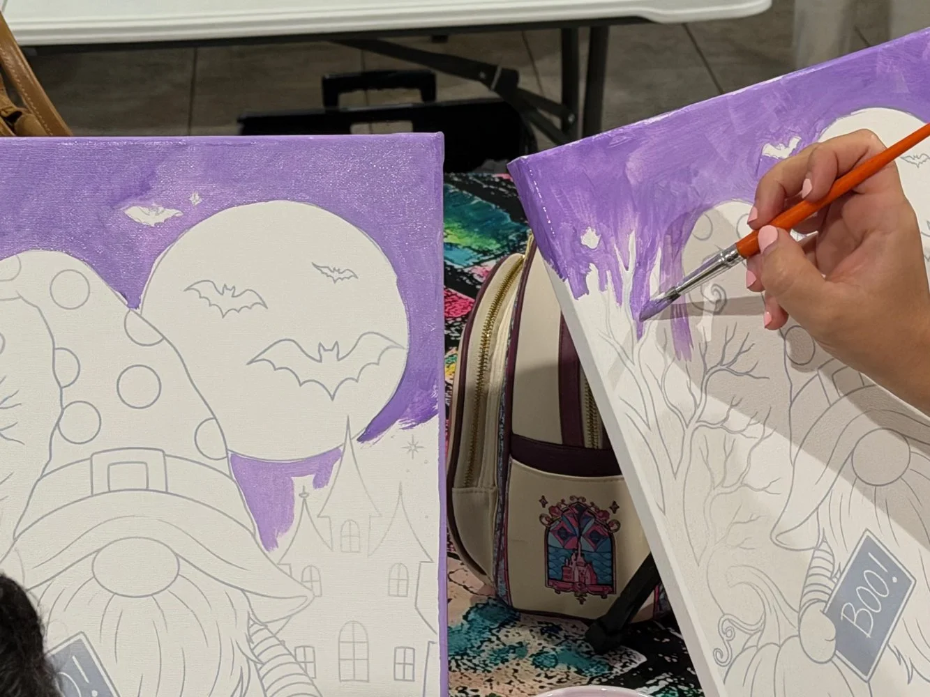 Person painting a Halloween-themed canvas with a purple background, bats, a gnome with a pointed hat, and a full moon.