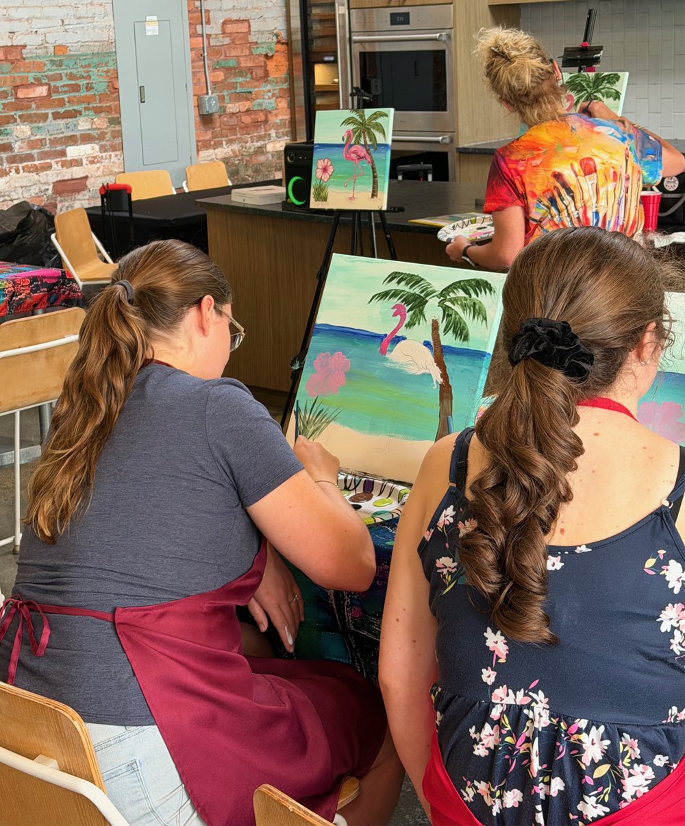 People painting tropical beach scenes with swans, palm trees, and pink flowers on easels in an indoor art class.