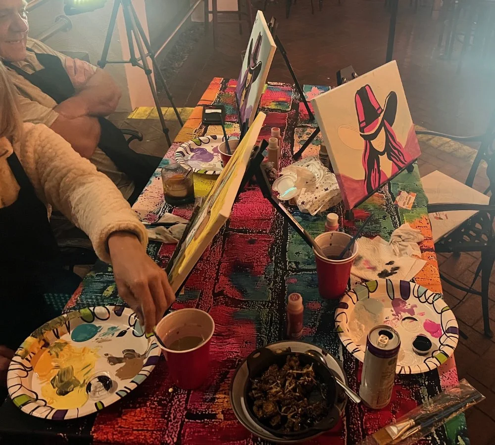 People painting canvases at a colorful table, with art supplies, plates, drinks, and napkins.