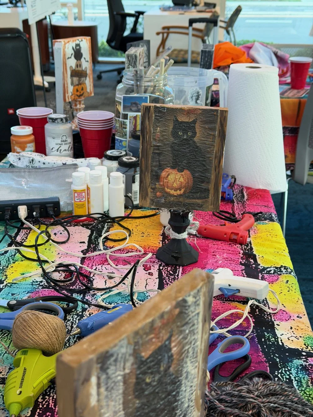 A tabletop covered with art supplies, including paint bottles, glue, scissors, paint brushes, and a lamp with a black cat and pumpkin painting. Red cups and paper towels are also on the table, with a colorful tablecloth underneath.