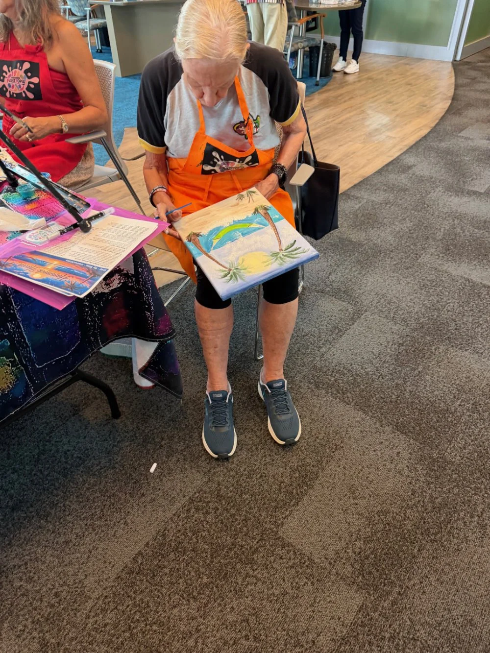 A woman sitting in a chair, painting a tropical beach scene on a canvas, wearing a striped t-shirt, orange apron, and athletic shoes in a room with a carpeted and wooden floor.