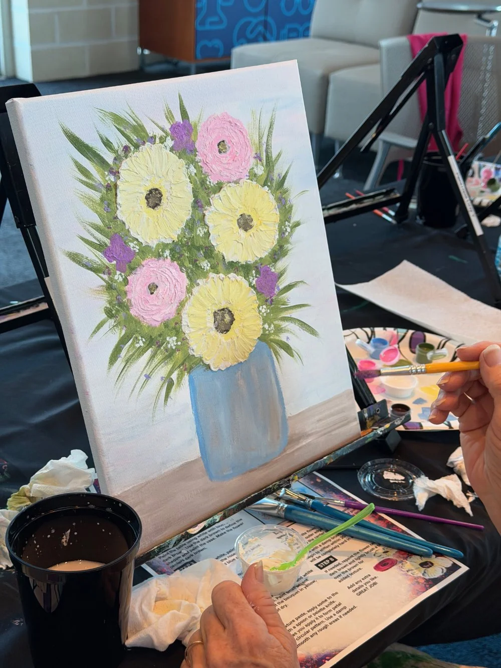 A person painting a floral still life on a canvas, depicting yellow and pink flowers in a blue vase, in an art class environment with painting supplies and a black table.